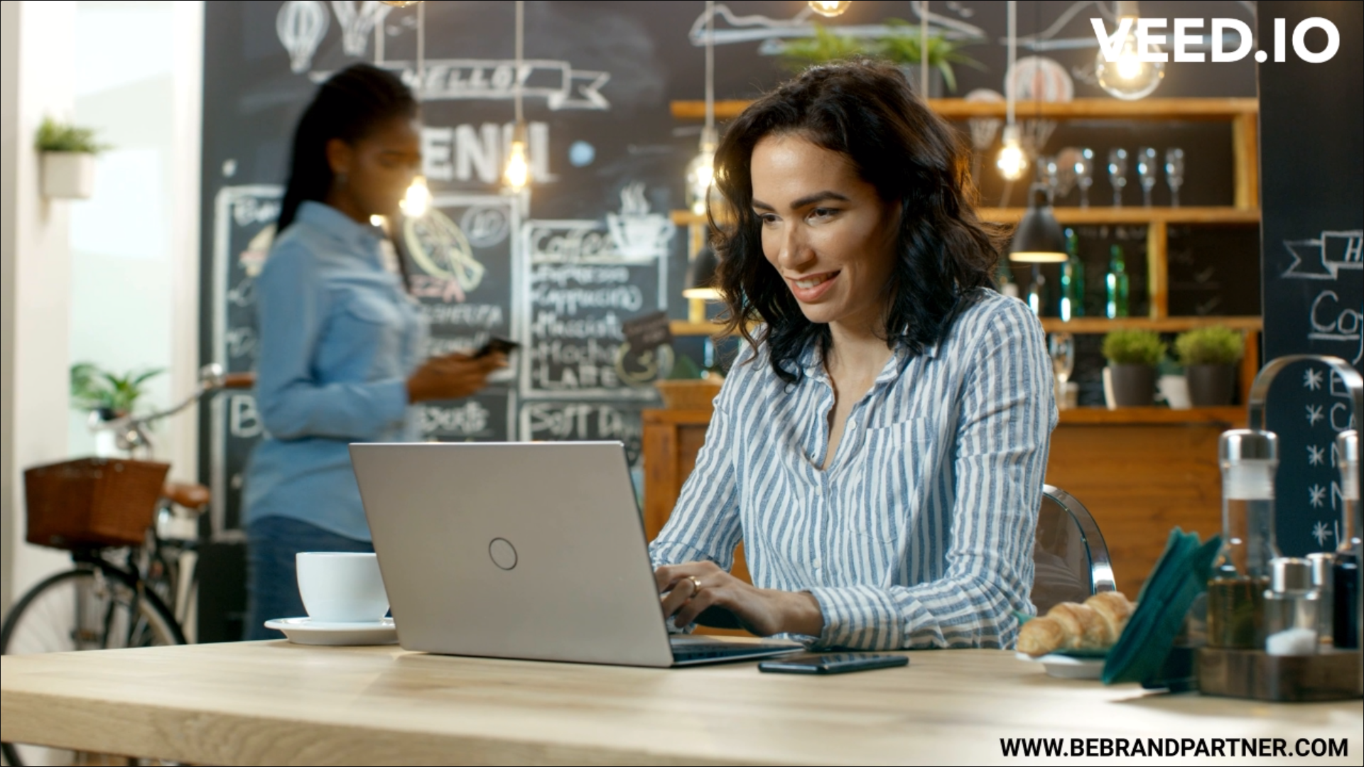 Woman working on laptop another woman in background using phone